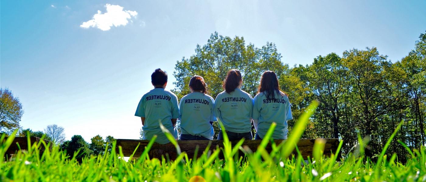 Four pharmacy students sit shoulder-to-shoulder on an outdoor bench wearing volunteer shirts
